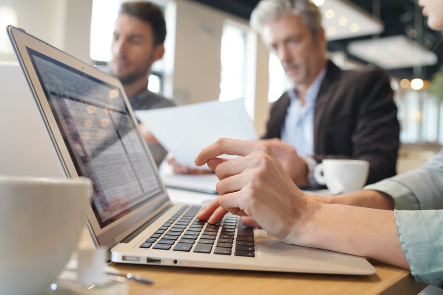 Hands typing on a laptop with colleagues talking in the background