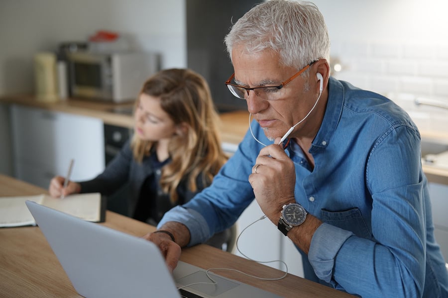 Man on a video call at a laptop while a child does homework nearby