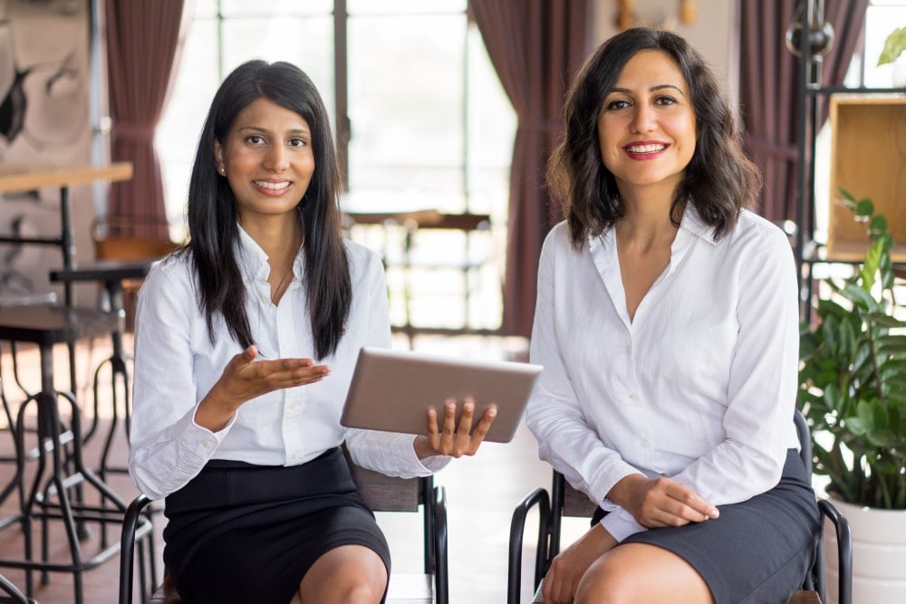 Two businesswomen seated in a café, one presenting on a tablet